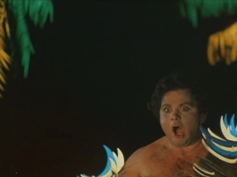 Salome (1972) movie still: medium — A dark environment populated by oversized tropical foliage and artificial palm leaves. A man appears from the…