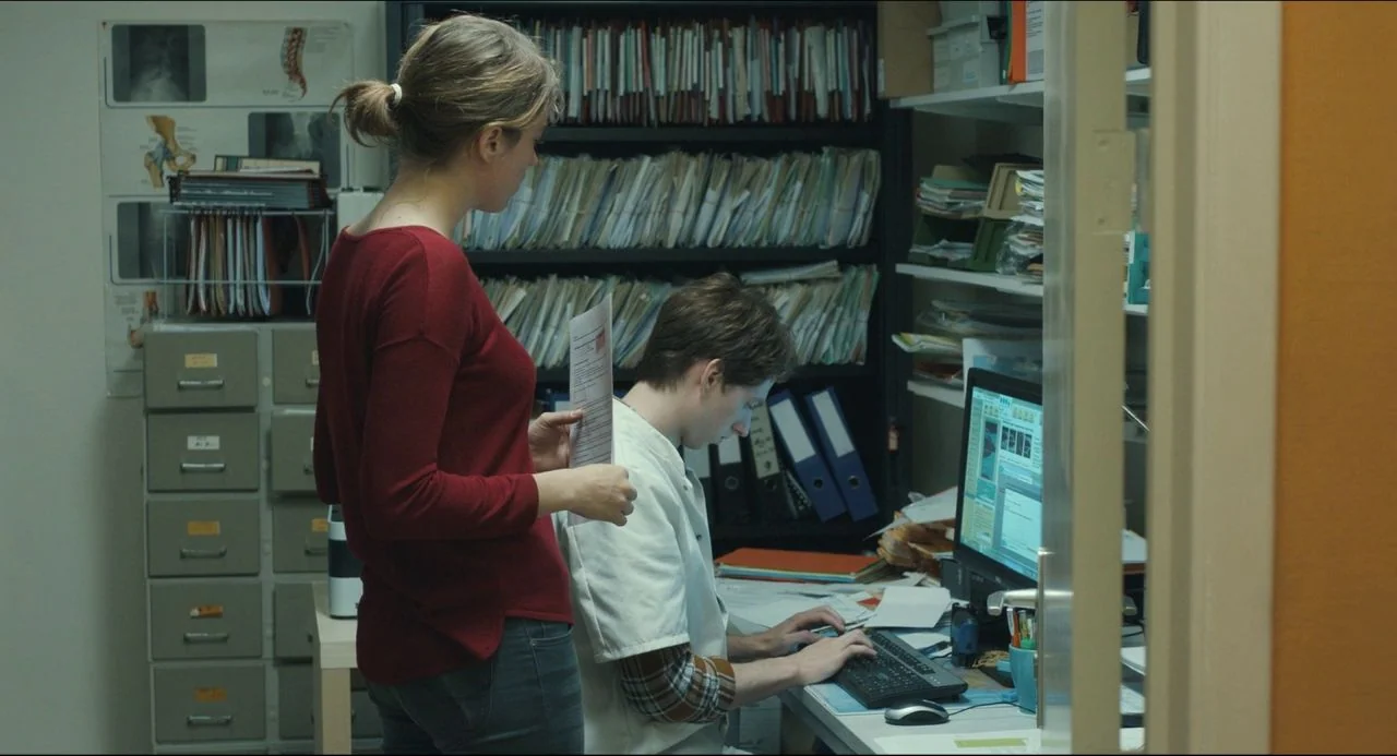 The Unknown Girl (2016) movie still: medium — A cluttered administrative work area is filled with shelves of binders and metal cabinets. A woman in…