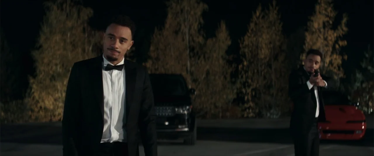 McWalter (2025) movie still: medium — A dark outdoor lot features trees in the background and two parked vehicles. One man in a tuxedo approaches…