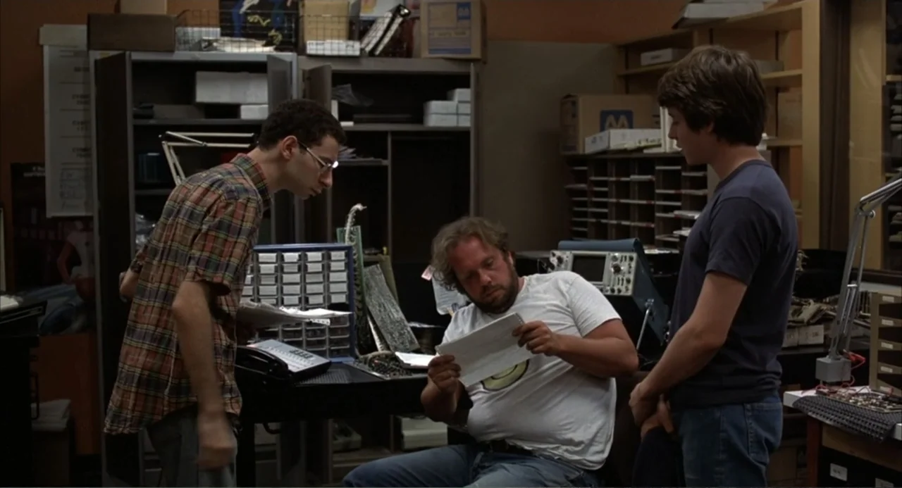 WarGames (1983) movie still: medium — A cluttered technical workshop contains shelves filled with boxes, electronic equipment, and components. Three…