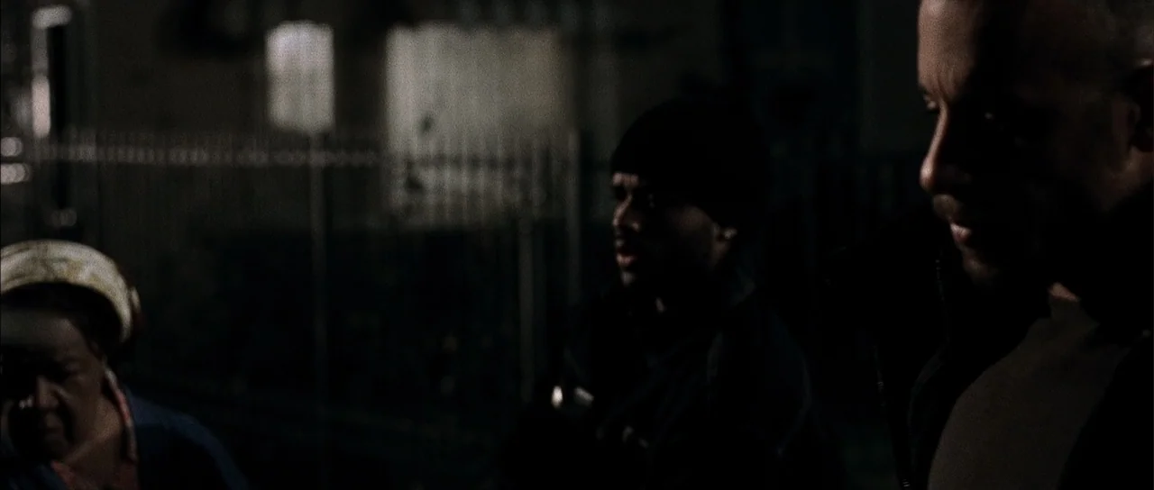 A Man Apart (2003) movie still: medium — An urban sidewalk area at night bordered by a metal chain-link fence. Three people stand together in…
