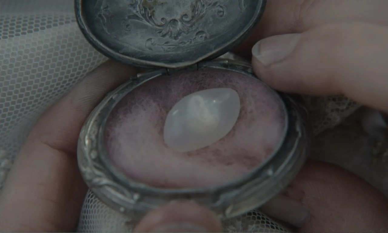 The Ugly Stepsister (2025) movie still: closeup — A close-up view shows a pair of hands holding an open vintage metal locket. A smooth, opaque white…