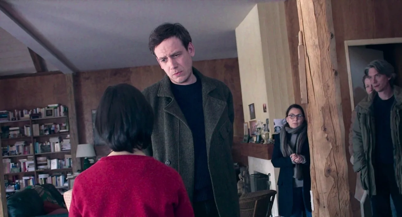 Anatomy of a Fall (2023) movie still: medium — A living room interior features a bookshelf filled with books and a wooden post. A man in a dark coat…