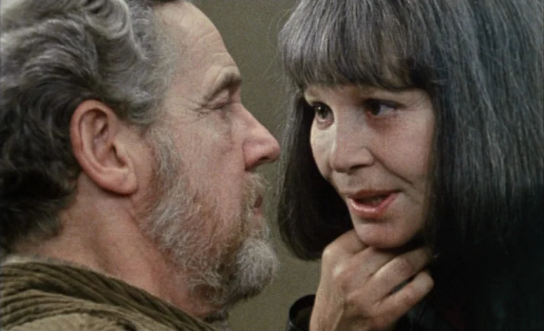After The Rehearsal (1984) movie still: closeup — Two individuals stand in a dimly lit space with a plain, neutral background. A woman holds the…