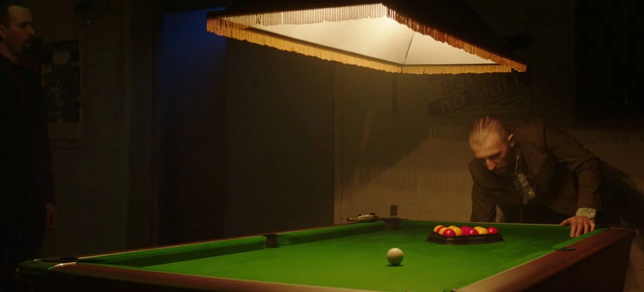 A Good Woman Is Hard To Find (2019) movie still: wide — A dim room contains a green pool table lit by a low-hanging fringe lamp. One man leans over…