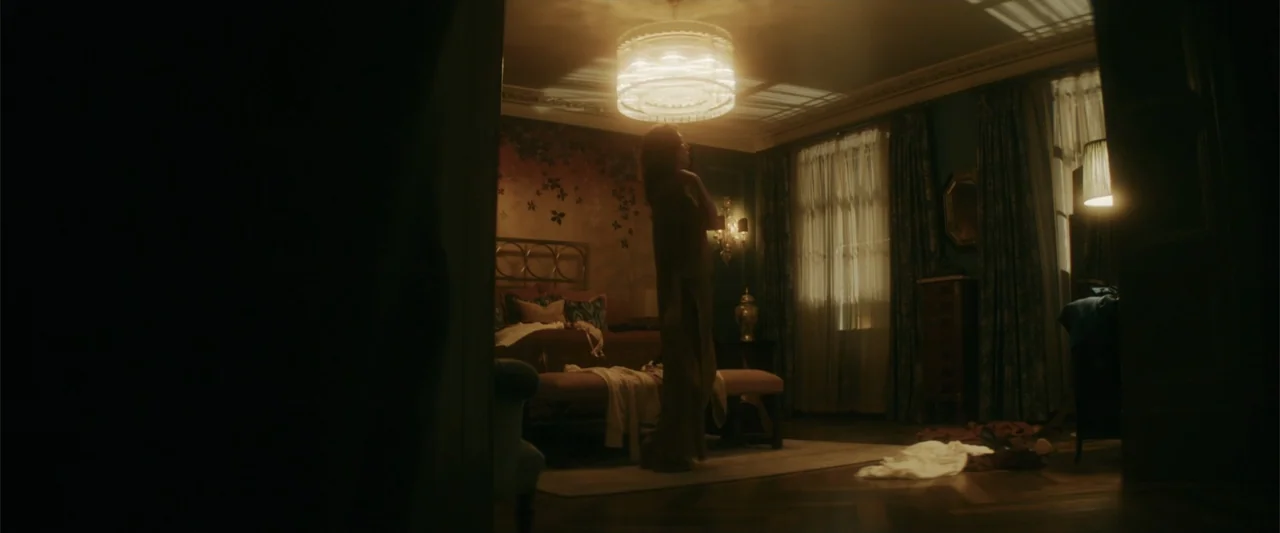 Black Bag (2025) movie still: fullbody — A dark, elegant bedroom features floral wallpaper, a neatly made bed, and a central chandelier. A woman…