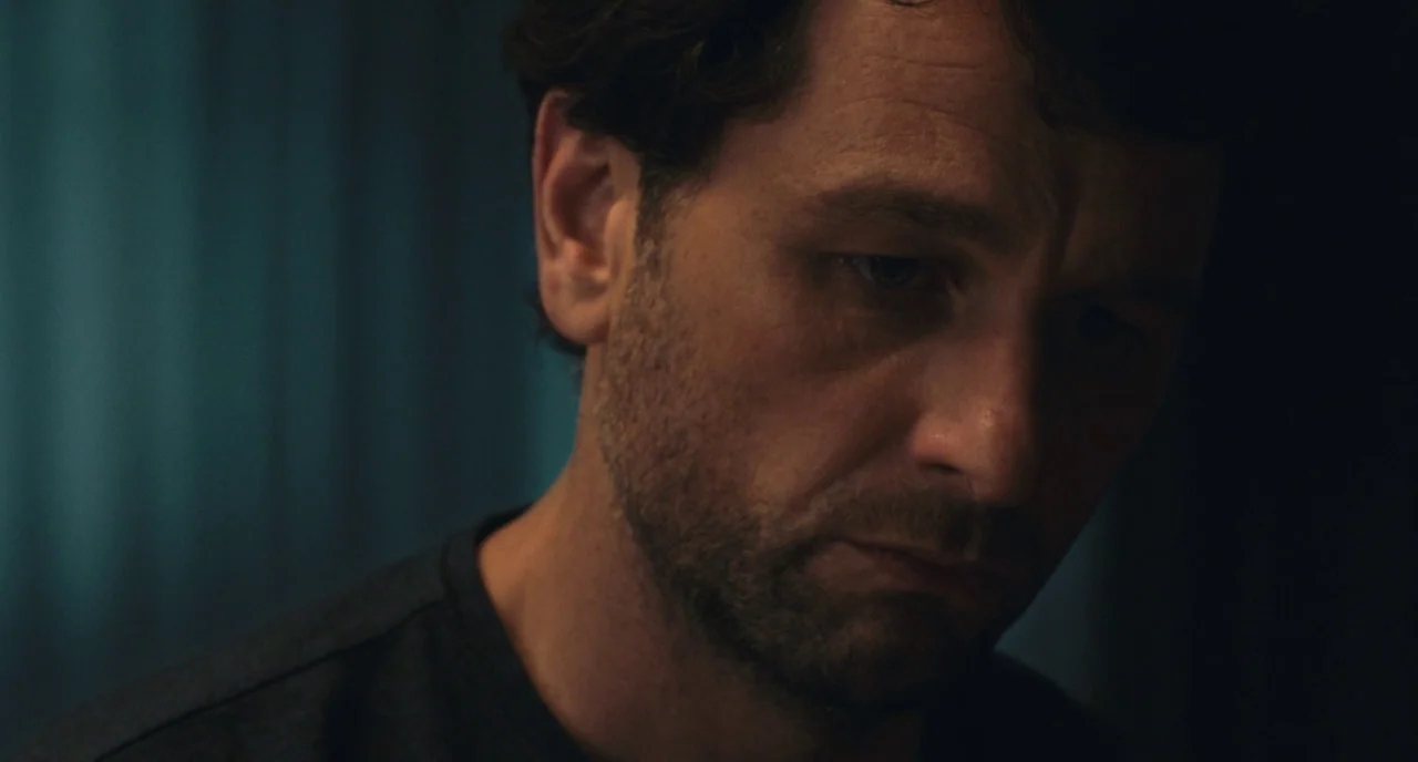 A Beautiful Day In The Neighborhood movie still: closeup — A man is positioned against a blurry, dark teal backdrop in a low-light room. He is…