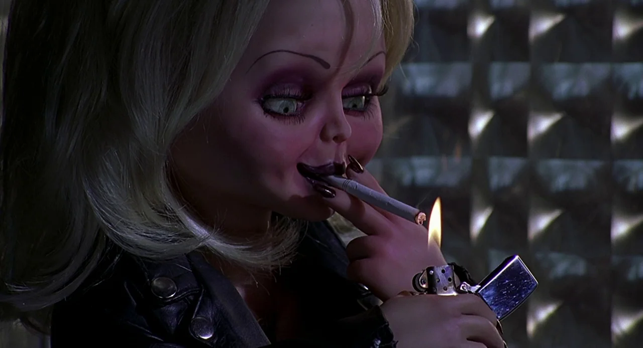 Bride of Chucky (1998) movie still: closeup — A blonde doll with heavy makeup sits in a dimly lit, dark space. The doll uses a metal cigarette…