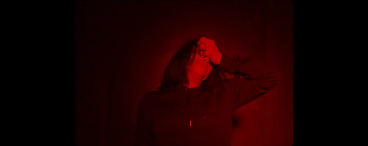 Lux Æterna (2019) movie still: medium — A person stands in a dimly lit, dark space illuminated by intense red light. The individual holds their hand…