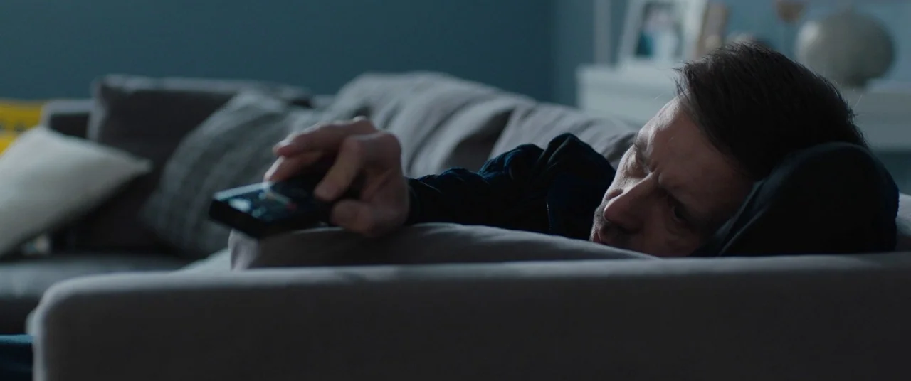 Journeyman (2017) movie still: medium — A man lies on a grey fabric sofa inside a dimly lit room. He holds a black remote control while resting his…