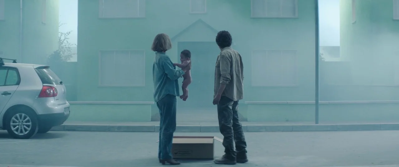 Vivarium (2019) movie still: wide — A quiet suburban street is blanketed in thick blue-tinted fog in front of houses. A man and a woman stand…