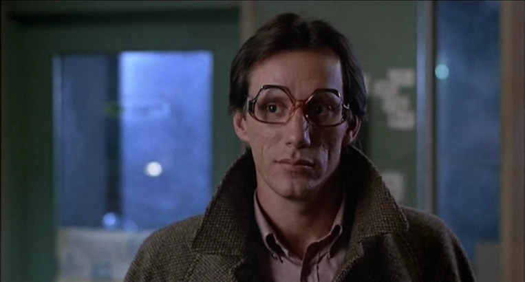 Videodrome (1983) movie still: medium — A dimly lit indoor corridor with muted green walls features a glass-paned door. A man in a brown tweed coat…