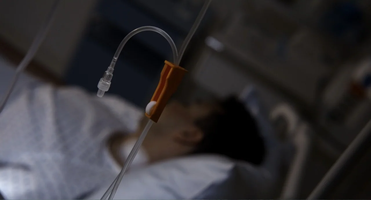 Gran Turismo (2023) movie still: closeup — A hospital room features medical apparatus in the foreground with a blurred person resting. A clear…