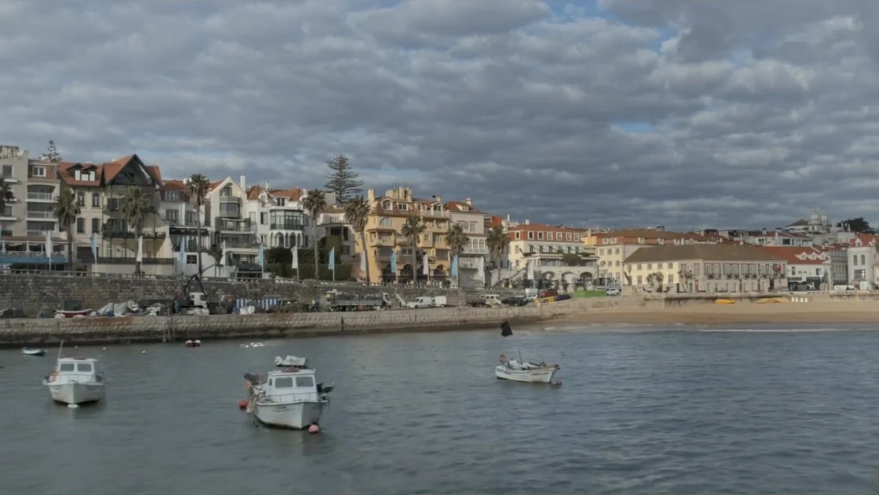 A Pinch of Portugal (2023) movie still: wide — A coastal town harbor features rows of multi-story buildings along a stone seawall. Several small…