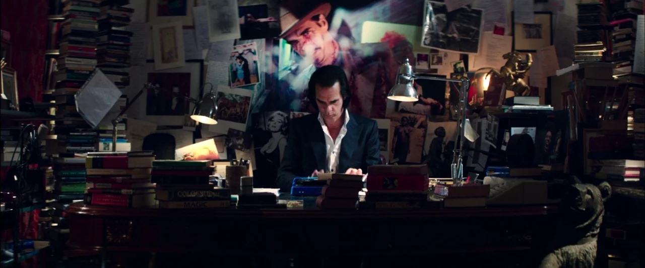 20,000 Days on Earth (2014) movie still: medium — A dark, cluttered room is filled with stacks of books and wall-mounted photographs. A man sits at…