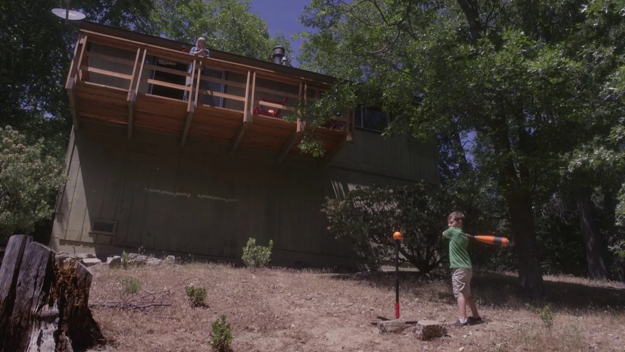 The Field Guide to Evil (2018) movie still: wide — A two-story green house stands on a hillside slope near a line of trees. A young boy practices…