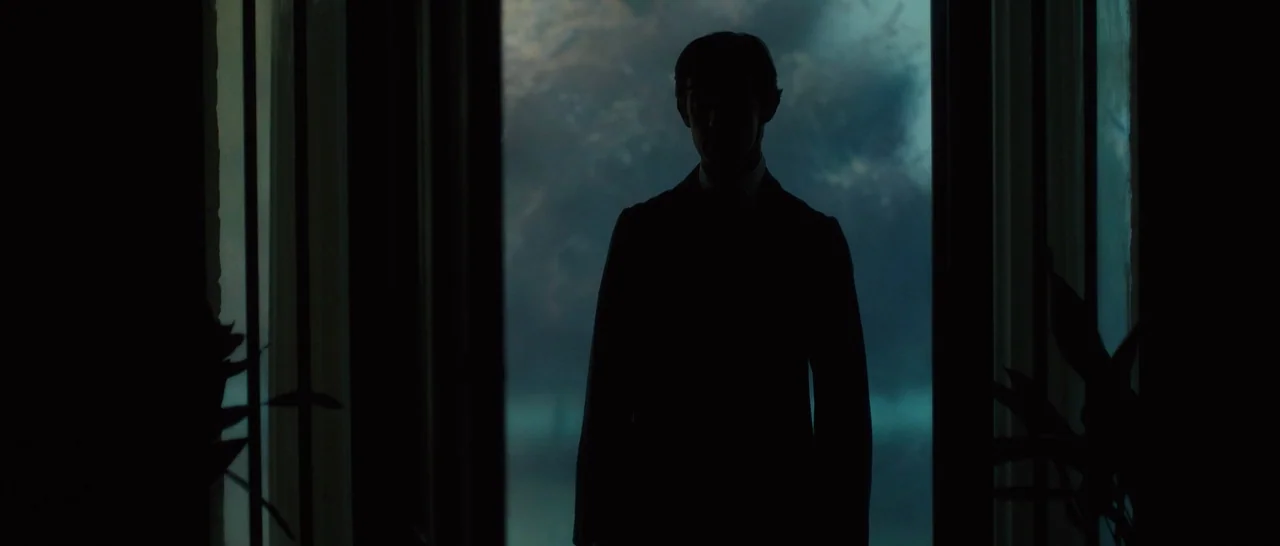 A Quiet Passion (2016) movie still: medium — A dark interior doorway frames a silhouette against a muted, foggy blue backdrop outside. A man in a…