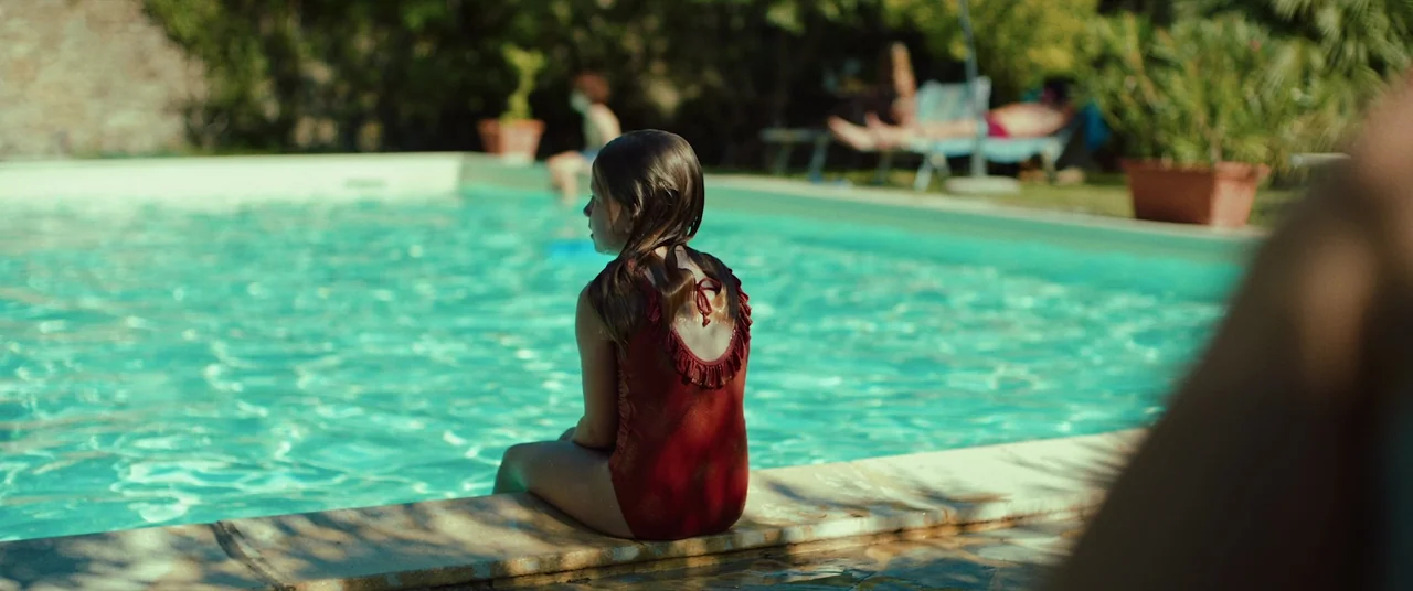Speak No Evil (2022) movie still: medium — A bright outdoor poolside area with clear turquoise water and nearby lounge chairs. A young girl in a red…