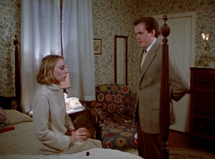 Desperate Living (1977) movie still: medium — A 1970s bedroom features floral wallpaper and a wooden bed frame near a window. A woman sits on the…