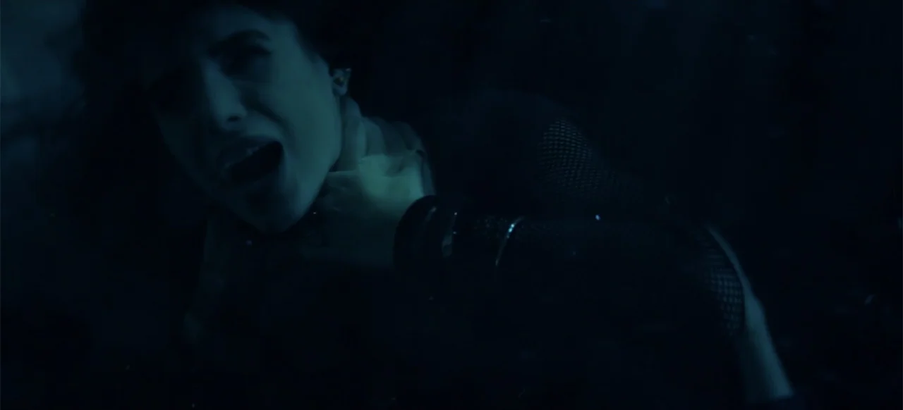 I Still See You (2018) movie still: closeup — A dark, underexposed interior space features a woman against a black background. The woman clutches…