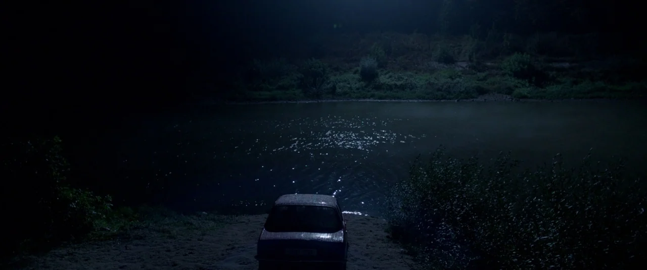 The Lure (2015) movie still: wide — A dark riverbank under a night sky with a single car facing the water. The opposite bank features dense…