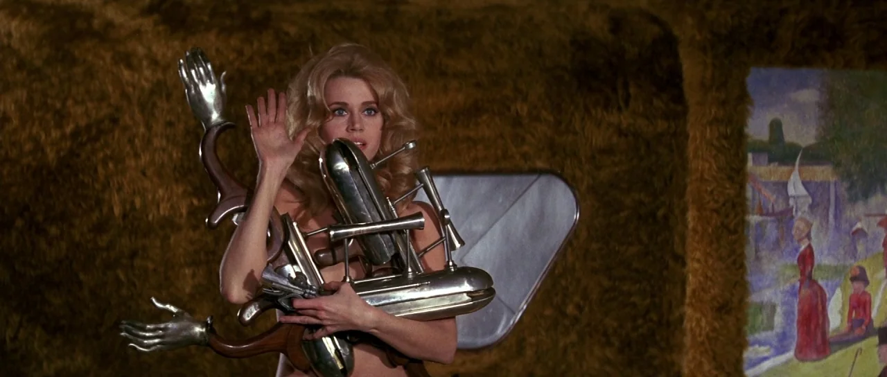 Barbarella (1968) movie still: medium — A woman stands in front of a wall covered in brown fur. She holds a complex metallic object featuring…