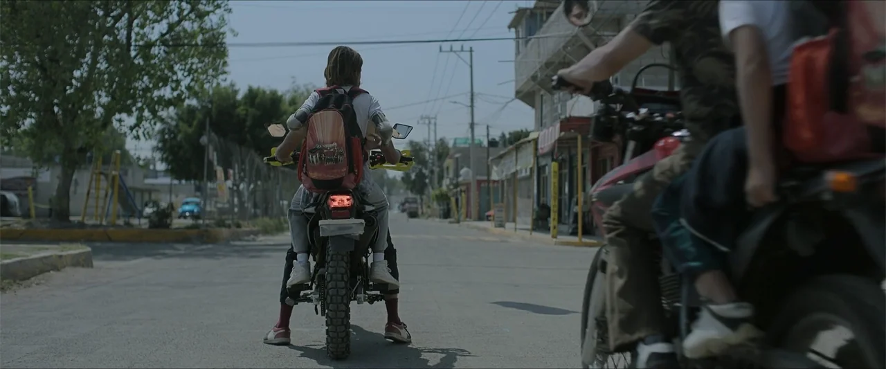 Radical (2023) movie still: medium — A quiet paved street in a small town features low-rise buildings and power lines. Two people on motorcycles are…