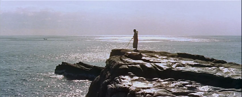 Kwaidan (1964) movie still: wide — A rocky headland juts into a wide, calm ocean under an overcast sky. A lone figure in robes stands on the edge…