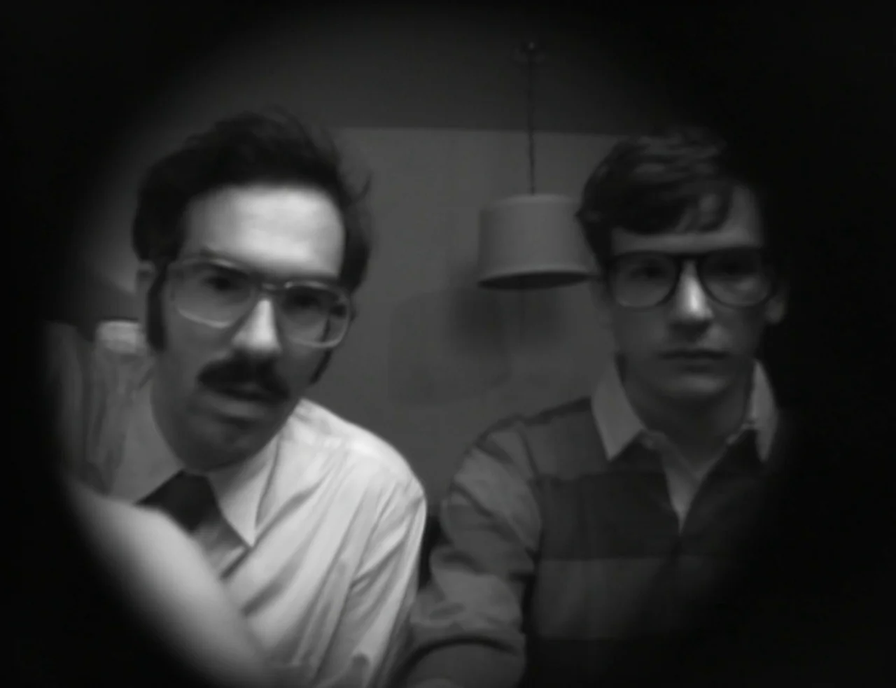 Computer Chess (2013) movie still: medium — Two men in glasses stare directly into the camera from a dimly lit space. A circular vignetted border…