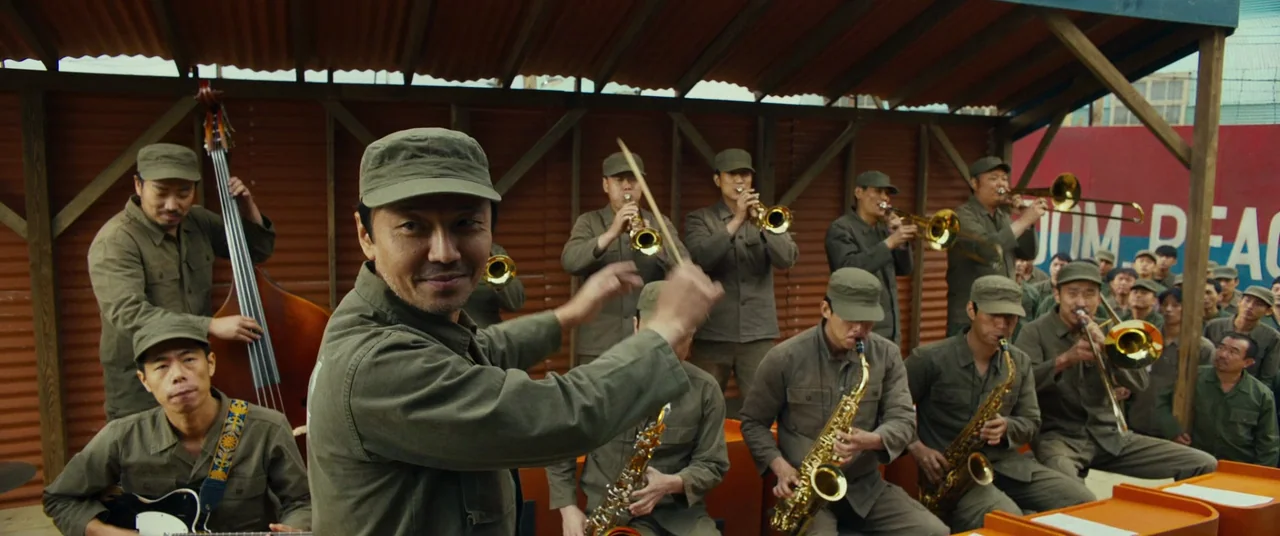Swing Kids (2018) movie still: medium — A group of men in military-style uniforms play musical instruments under a corrugated roof. The band leader…