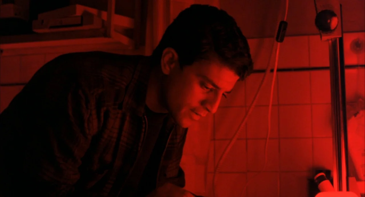 The Good Thief (2002) movie still: medium — A darkroom features tiled walls and a bright red safelight fixture on the right. A young man stands in…