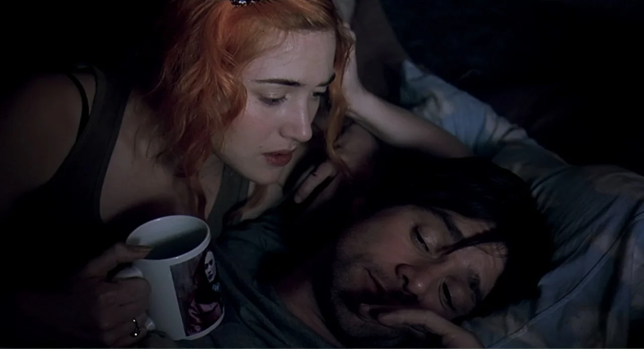 Eternal Sunshine of the Spotless Mind (2004) movie still: closeup — A dimly lit bedroom features two people resting together in a bed. A woman with…