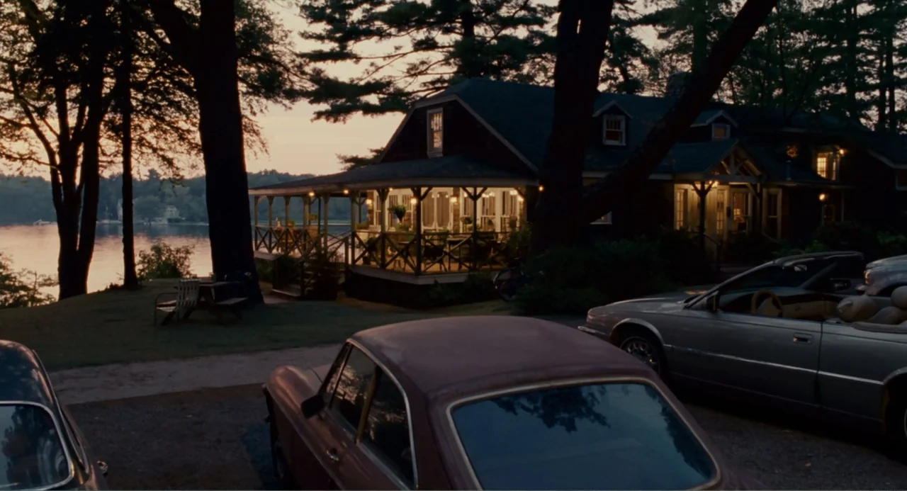 Grown Ups (2010) movie still: establishing — A rustic wooden house sits on the edge of a lake at dusk. Several vehicles are parked in the gravel…