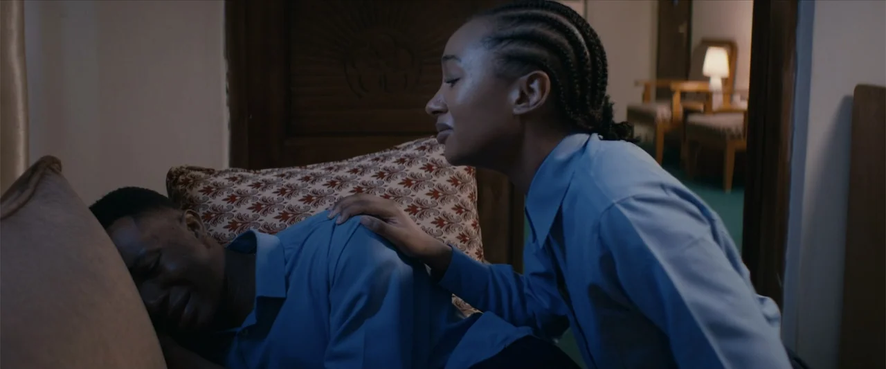 Ms. Kanyin (2025) movie still: medium — A hotel room with a patterned bed pillow and a lamp in the distance. A woman kneels beside a man reclining…