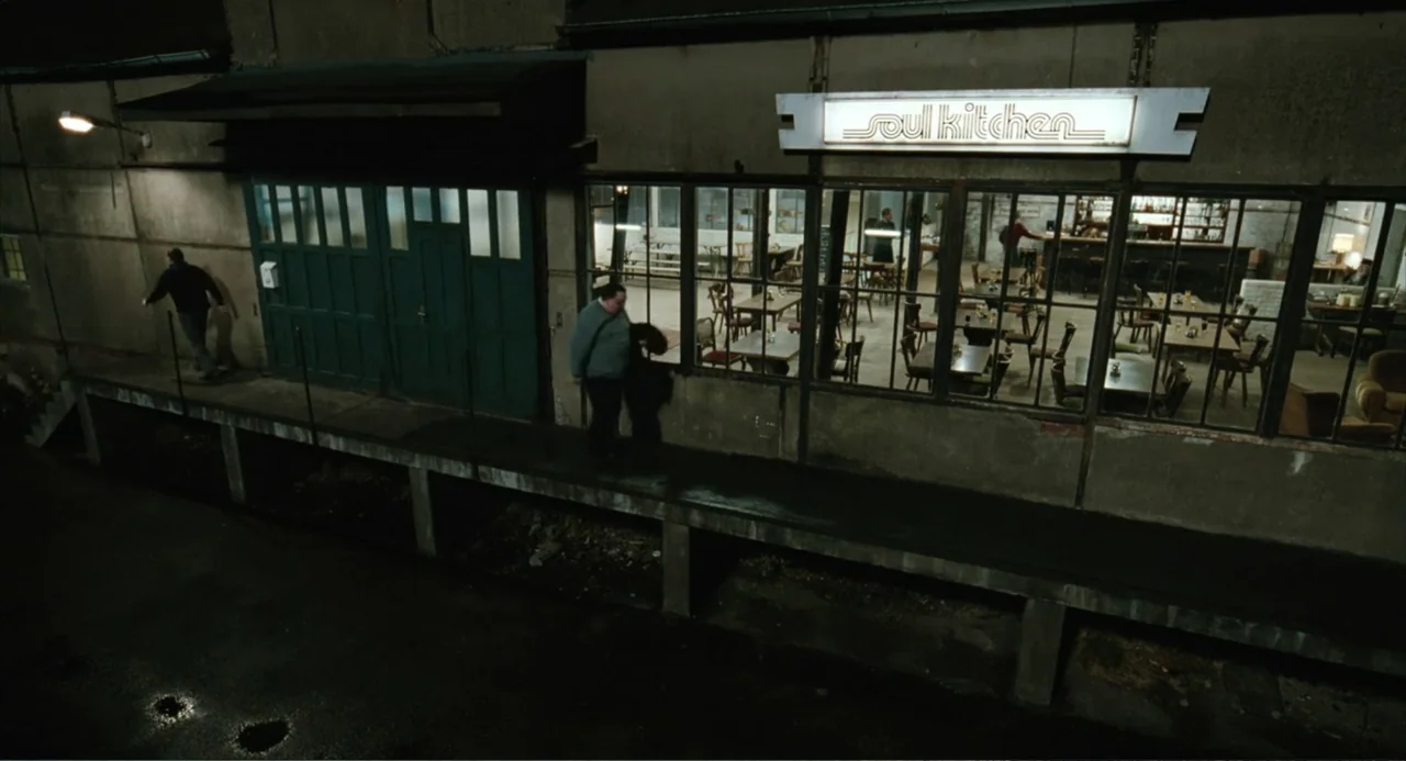 Soul Kitchen (2009) movie still: wide — An exterior view of a restaurant named Soul Kitchen at night with a loading dock. A man walks along the…