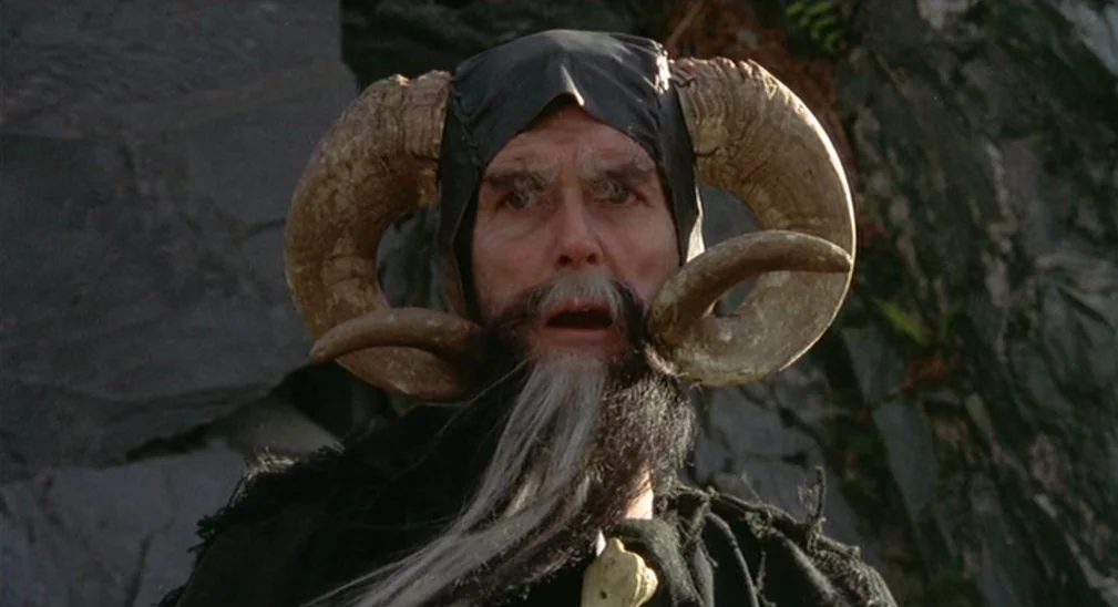 Monty Python and the Holy Grail (1975) movie still: closeup — A rugged rock face provides a natural backdrop for a fantasy figure. An elderly man…