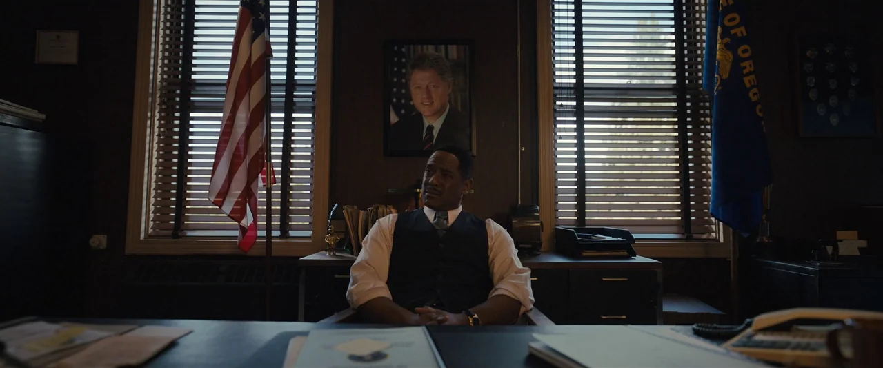 Longlegs (2024) movie still: medium — A dimly lit office features dark wood paneling, American and Oregon flags, and window blinds. A man sits in a…