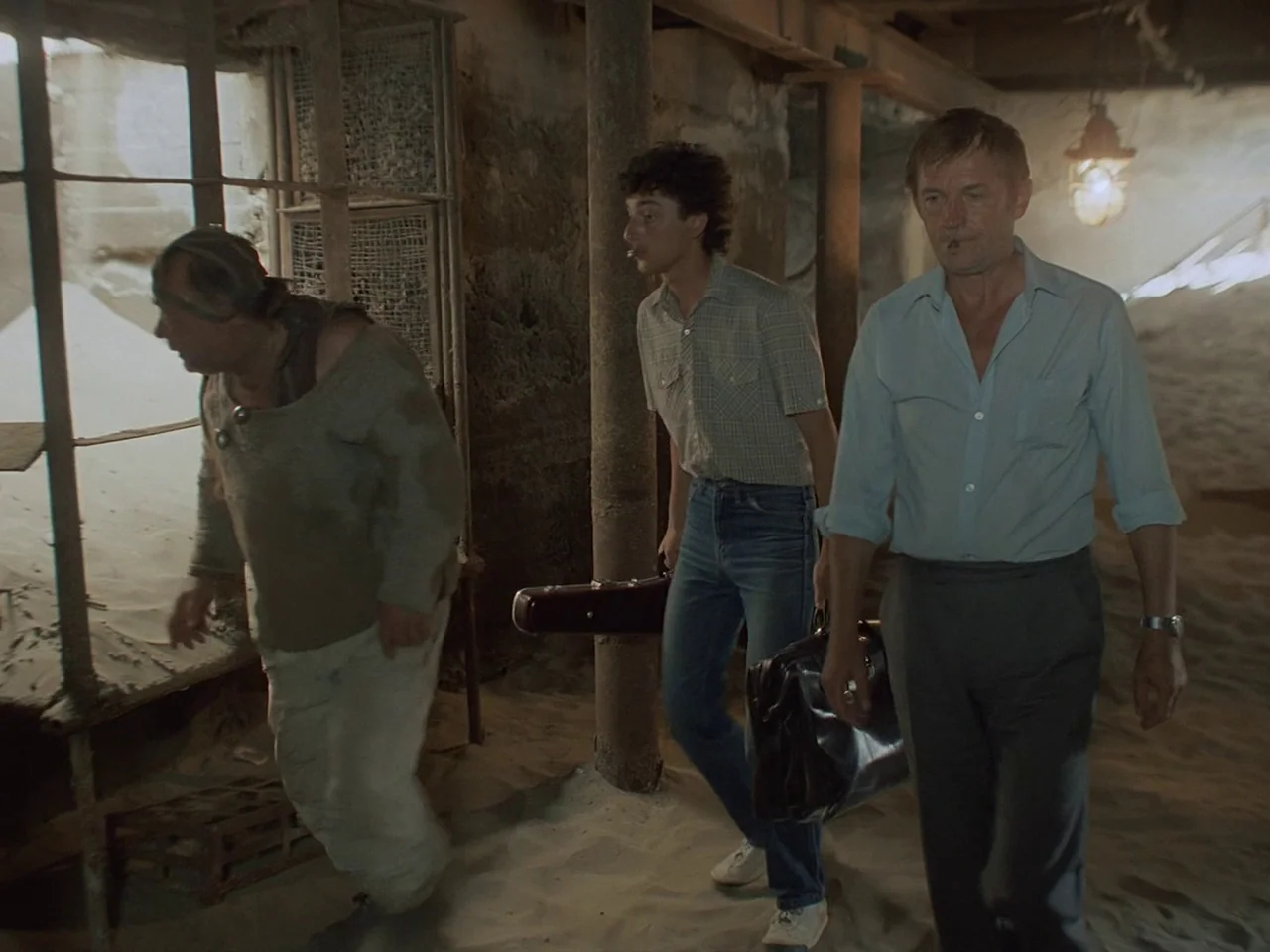 Kin-dza-dza! (1986) movie still: fullbody — An interior space with a floor covered in thick sand and concrete pillars. Three men walk together…