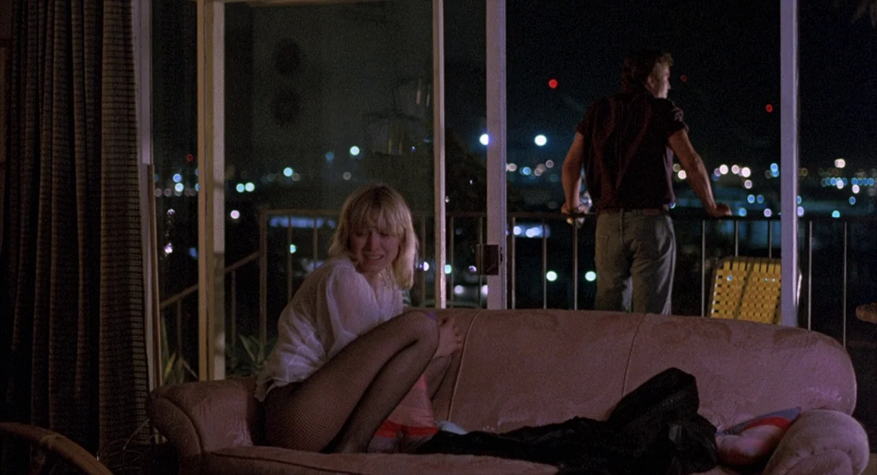 To Live and Die in L.A. (1985) movie still: medium — A dimly lit room with a pink sofa facing a large sliding glass door. The man stands on the…