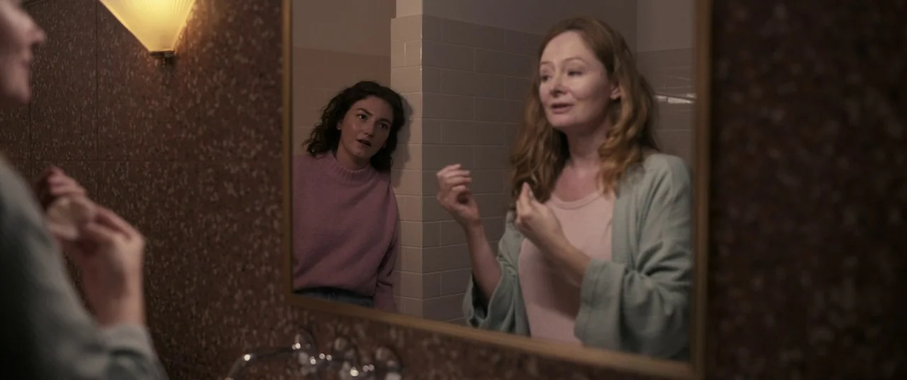Talk To Me (2022) movie still: medium — A tiled bathroom features a large wall mirror with a gold-toned frame. One woman looks into the mirror while…