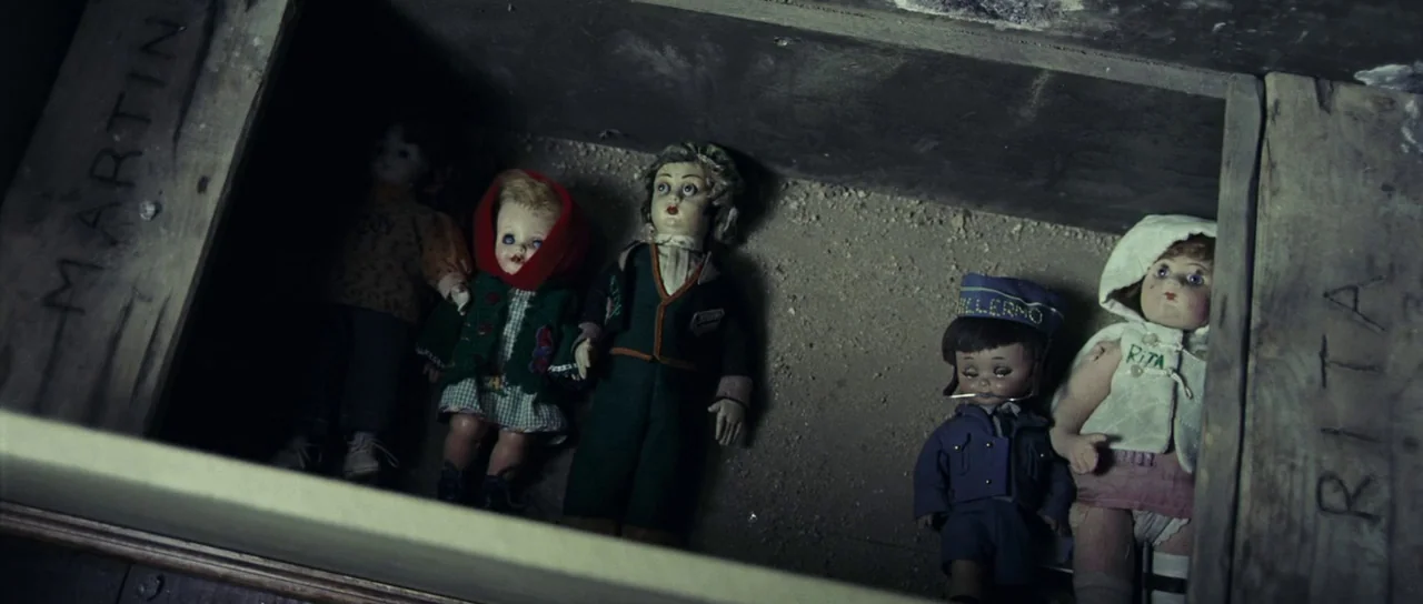 The Orphanage (2007) movie still: medium — A dark wooden crate holds five assorted vintage dolls resting on a dusty surface. The edges of the crate…
