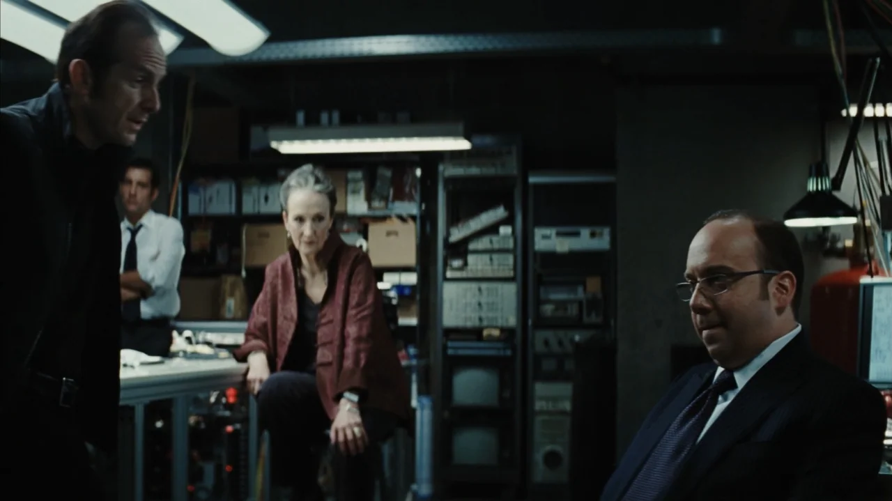 Duplicity (2009) movie still: medium — A dimly lit technical room contains computer racks, cables, and various electronic equipment. Three people…