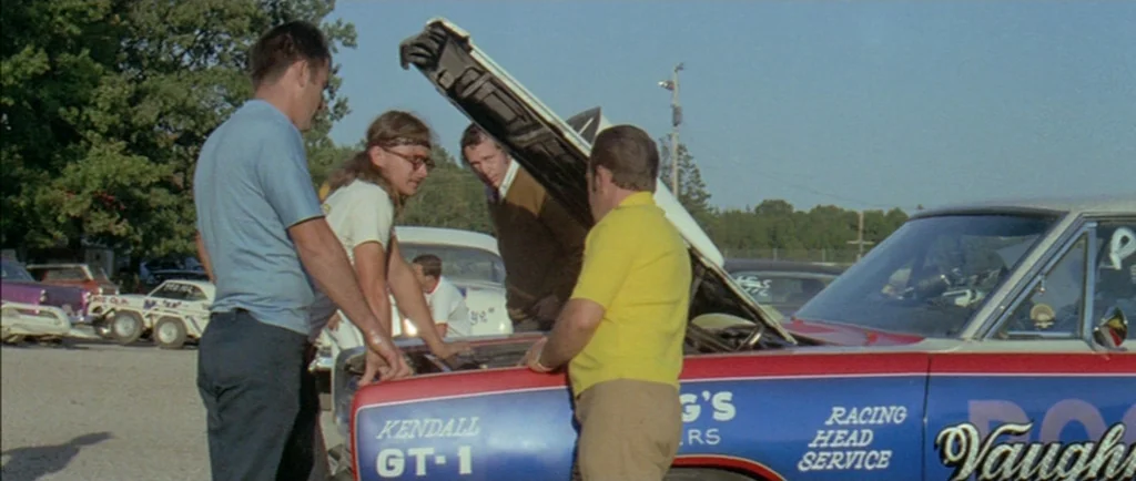 Two Lane Blacktop (1971) movie still: fullbody — A group of men gather around a race car with its hood propped open. The scene takes place on a…
