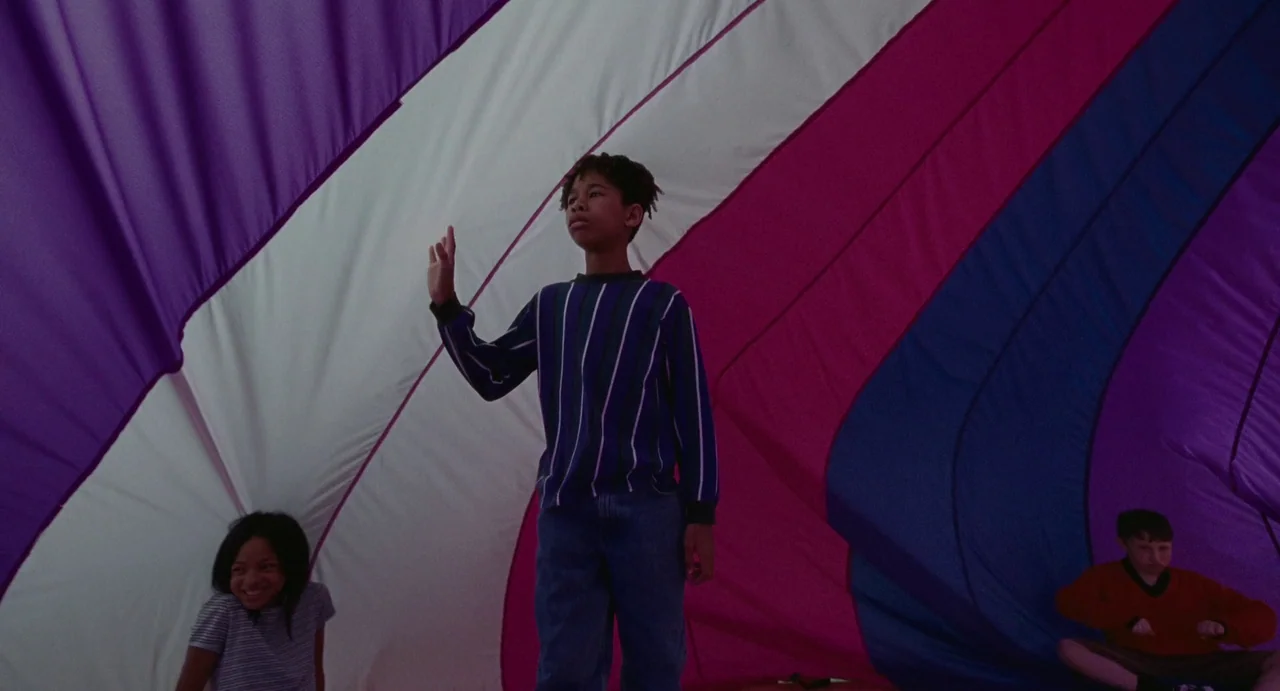 I Saw The TV Glow (2024) movie still: fullbody — Children play inside the canopy of a giant multi-colored gymnasium parachute. Three children are…