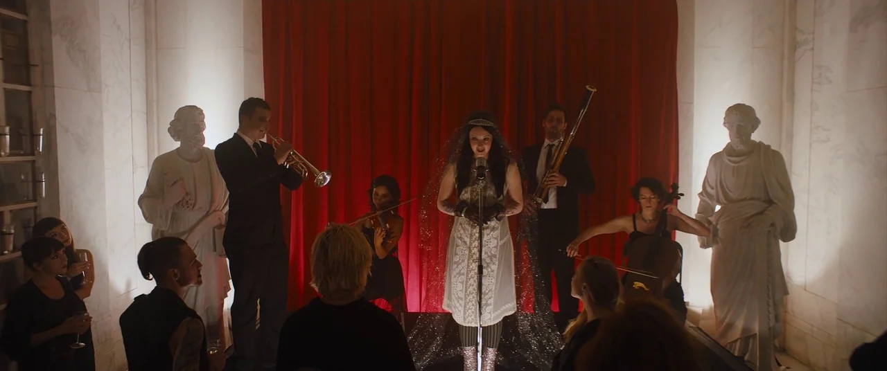 Under The Silver Lake (2018) movie still: wide — A group of musicians performs in front of a red curtain between two large statues. A woman stands…