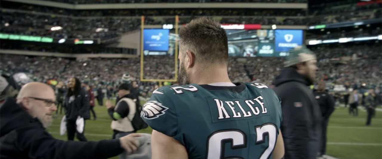 Kelce (2023) movie still: medium — A crowded football field at night with large bright stadium lights and digital displays. A male athlete in a…
