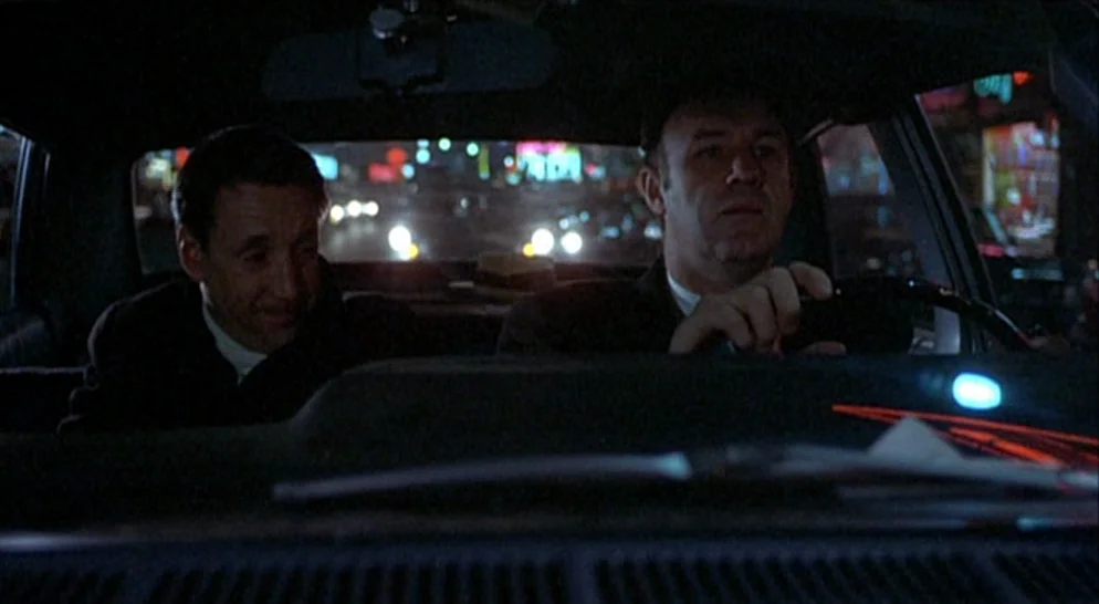 The French Connection (1971) movie still: medium — An interior shot of a 1970s sedan at night on a city street. Two men sit in the front seats while…