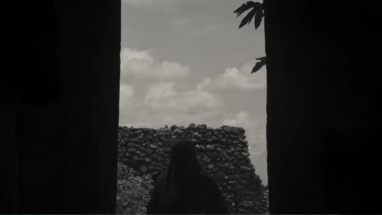 Faya Dayi (2021) movie still: medium — A dark stone-walled passage frames an exterior view of a low rock wall. A silhouette of a person in a hooded…