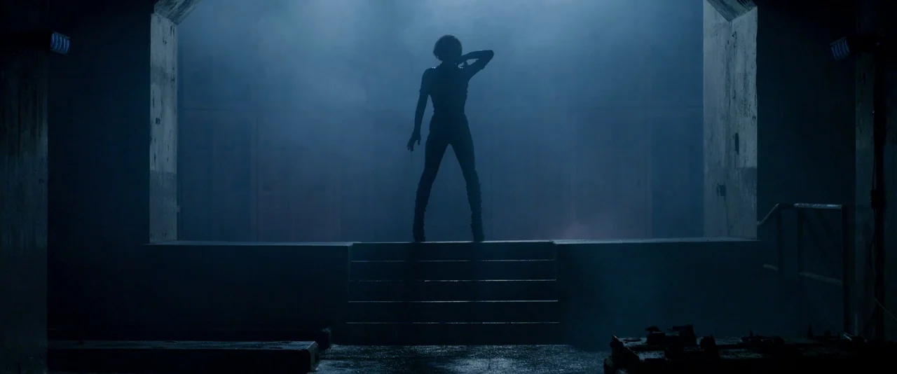 Resident Evil: Retribution (2012) movie still: wide — A dark concrete chamber with a raised platform illuminated by a hazy blue light. A lone female…