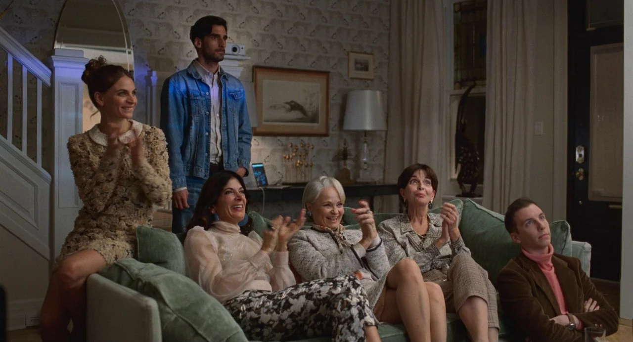 Matthias & Maxime (2019) movie still: wide — A well-lit living room contains a sage green sofa and patterned wallpaper. Six people stand or sit on…